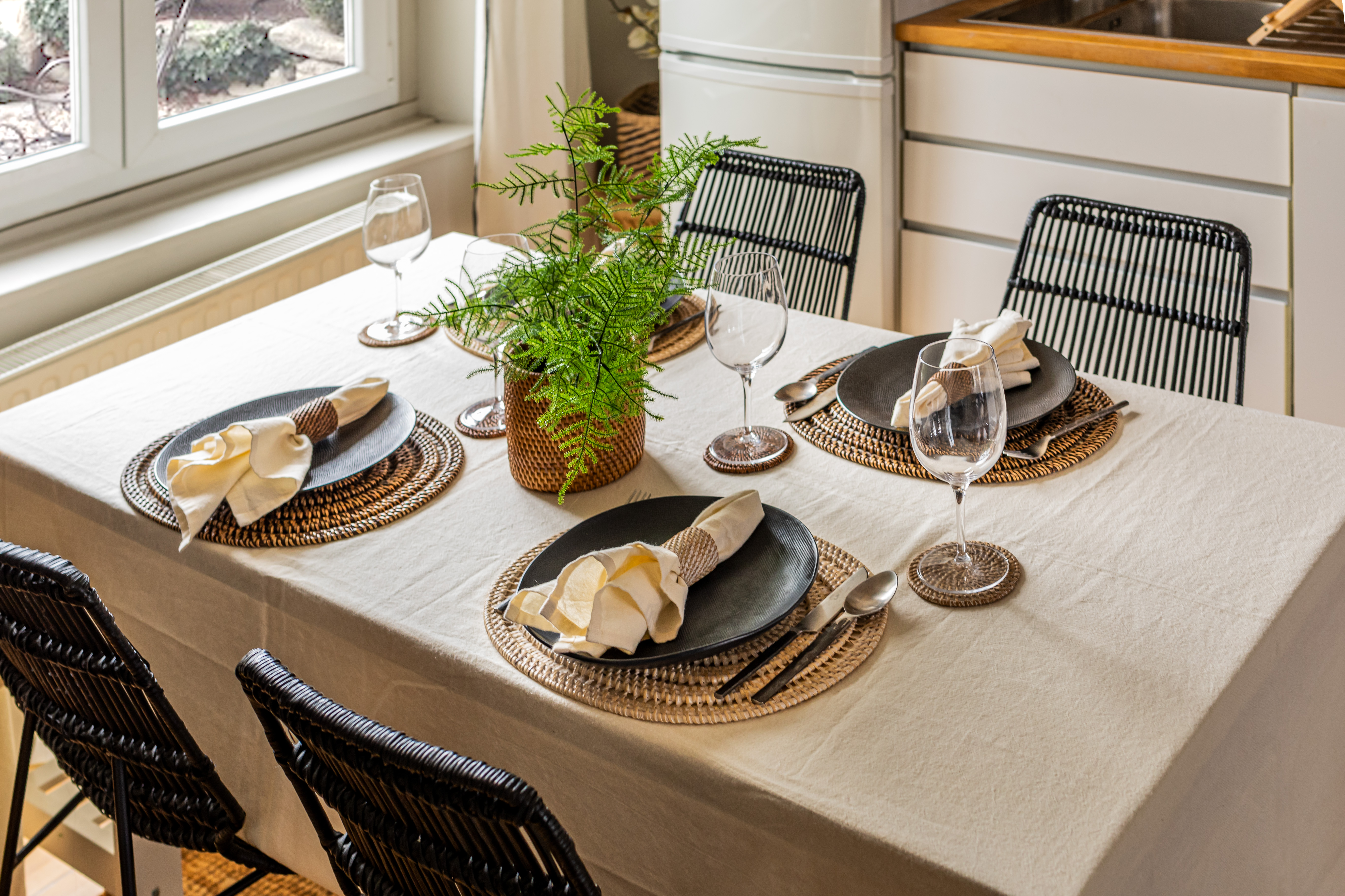 Een gedekte tafel met een tafelkleed waarop ronde placemats, glazen onderzetters en rotan servetringen liggen.